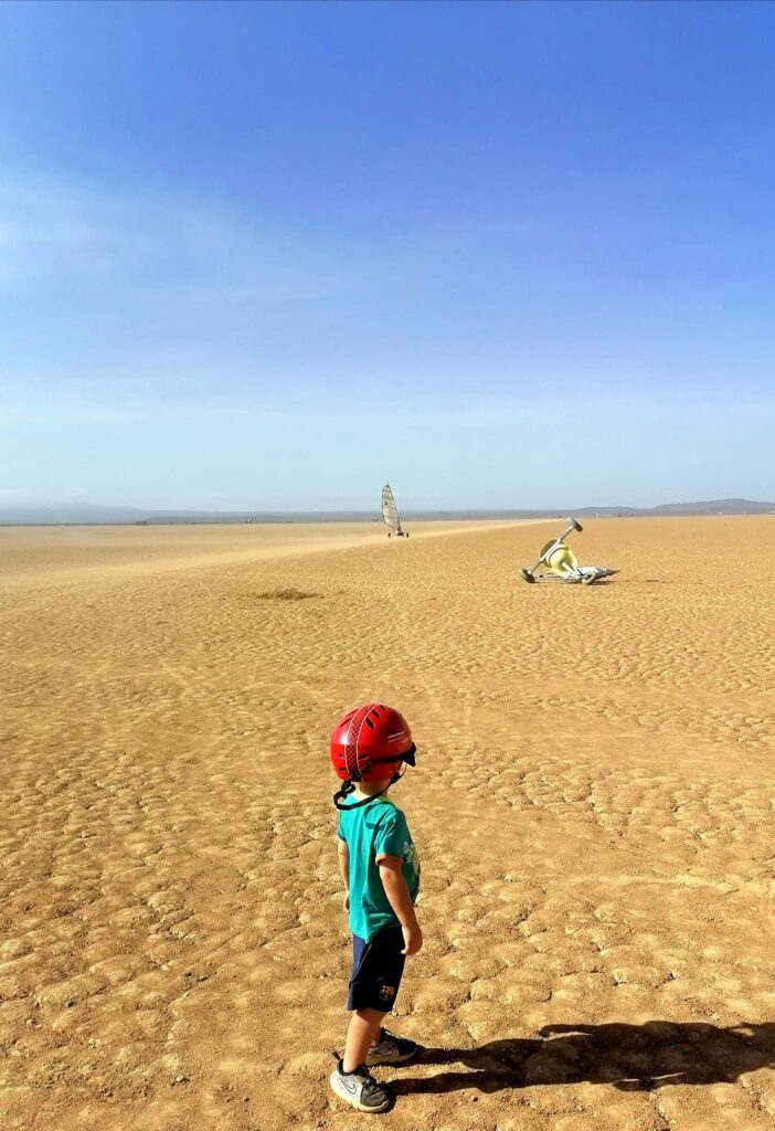 Un enfant en casque rouge observe des chars à voile glissant sur les vastes étendues sablonneuses du désert djiboutien sous un ciel bleu dégagé. Une image qui capture l'esprit d'aventure et de liberté offert par le Djibouti Land Yachting Club.