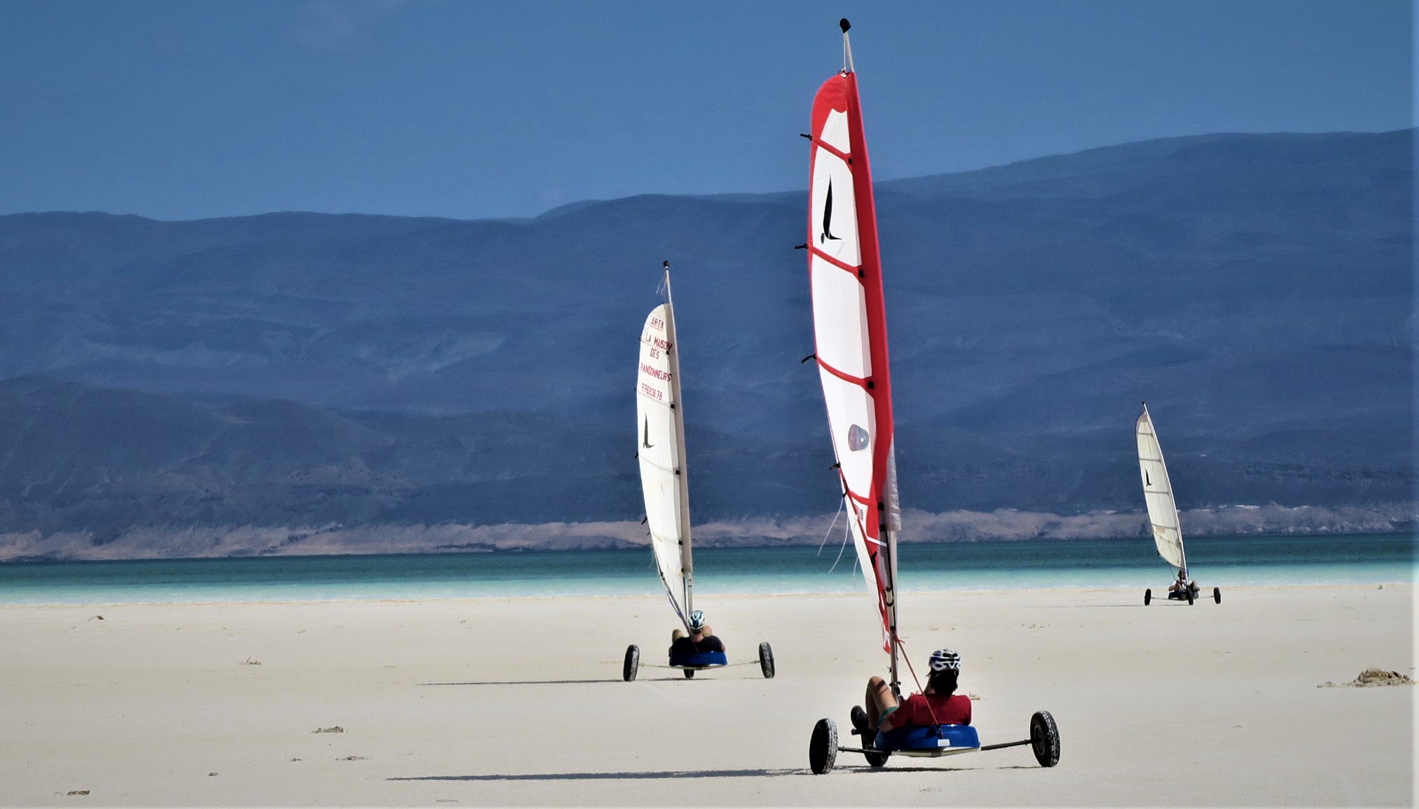 Des chars à voile traversent une plage de sable blanc avec en toile de fond des eaux turquoise et des montagnes majestueuses.