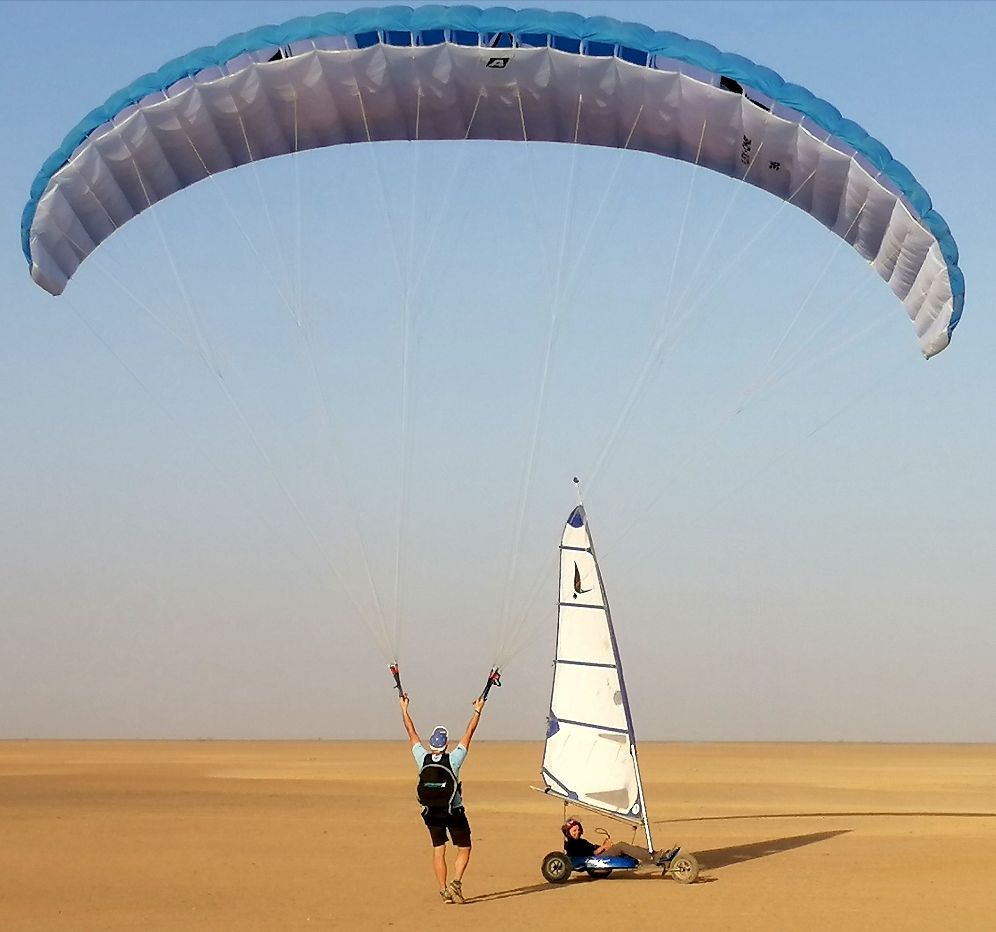 Un homme en parapente s'élève au-dessus du désert, tandis qu'un char à voile avance sur le sable doré.